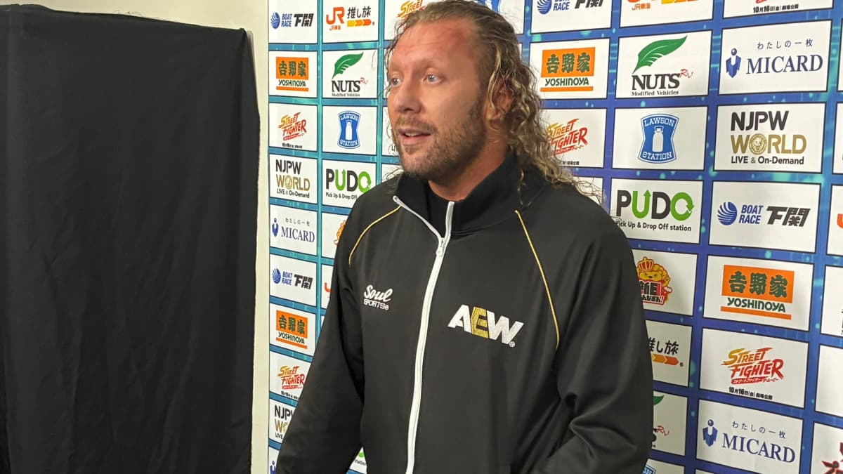 Kenny Omega in an AEW tracksuit, speaking backstage at an NJPW event