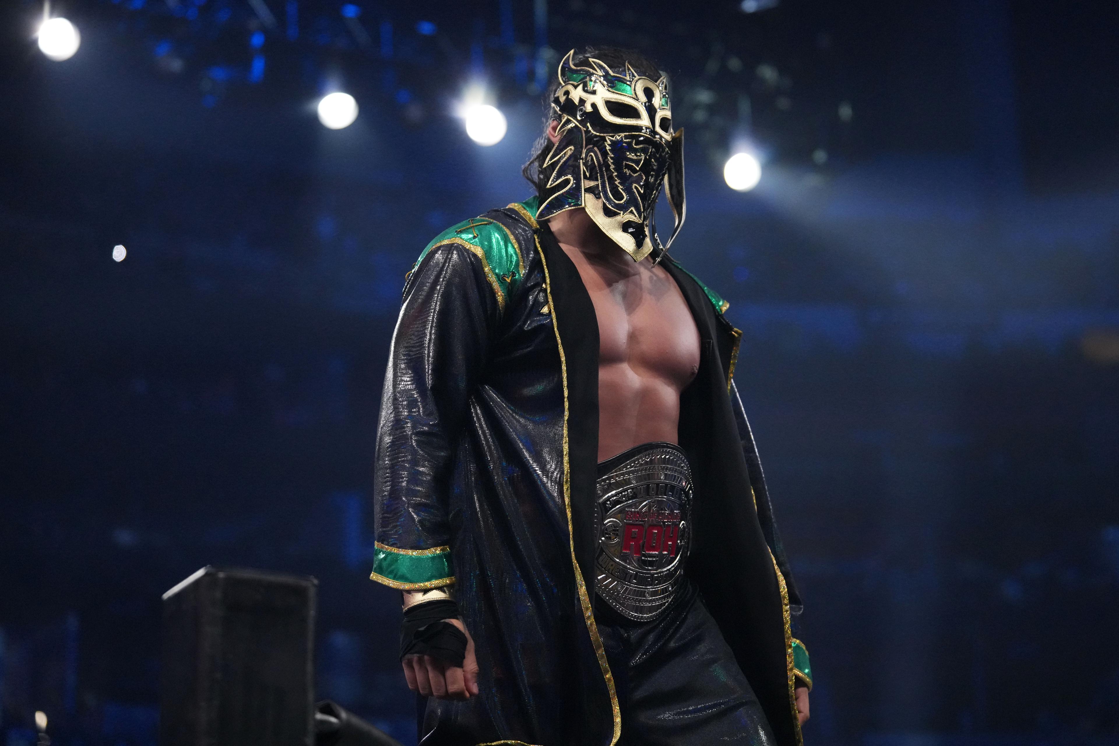 Bandido with a Super Dragon-esque mask and and a green jacket with the ROH World Title around his waist while standing on a turnbuckle