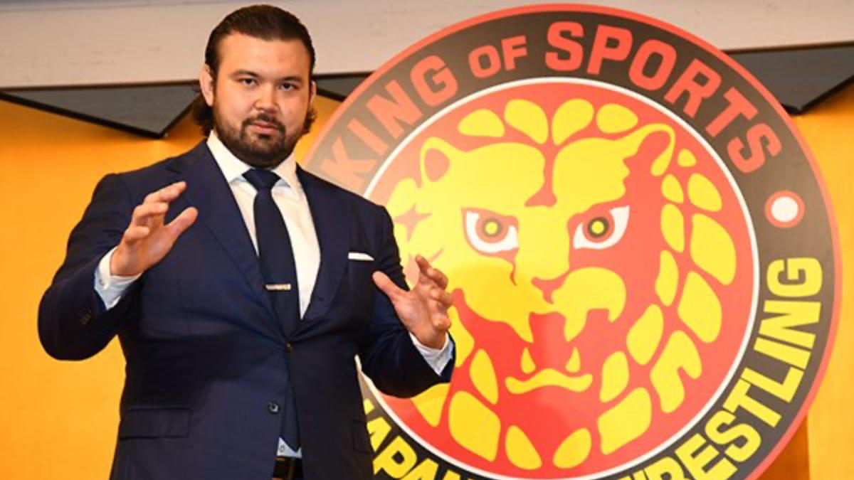 Aaron Wolf standing in front of the NJPW logo while wearing a suit