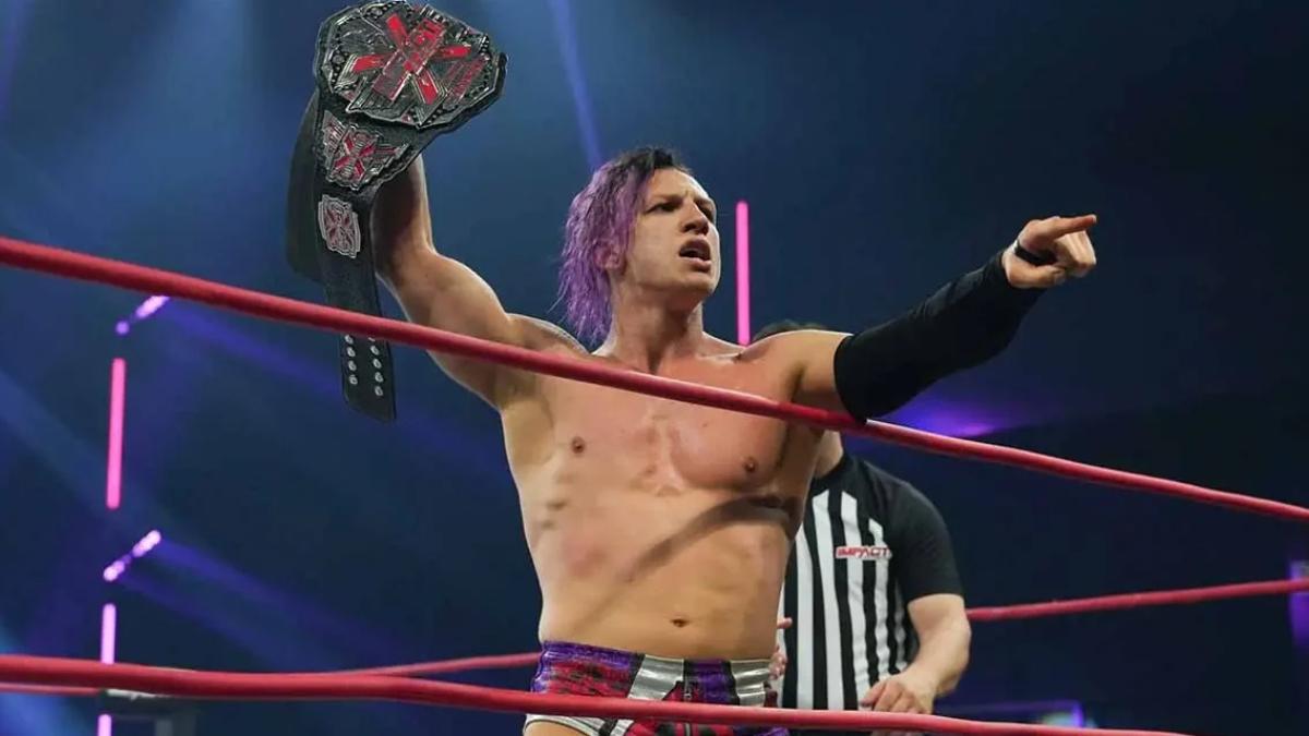 Ace Austin inside a wrestling ring holds up the X Division Championship with one hand, and points to the audience with the other