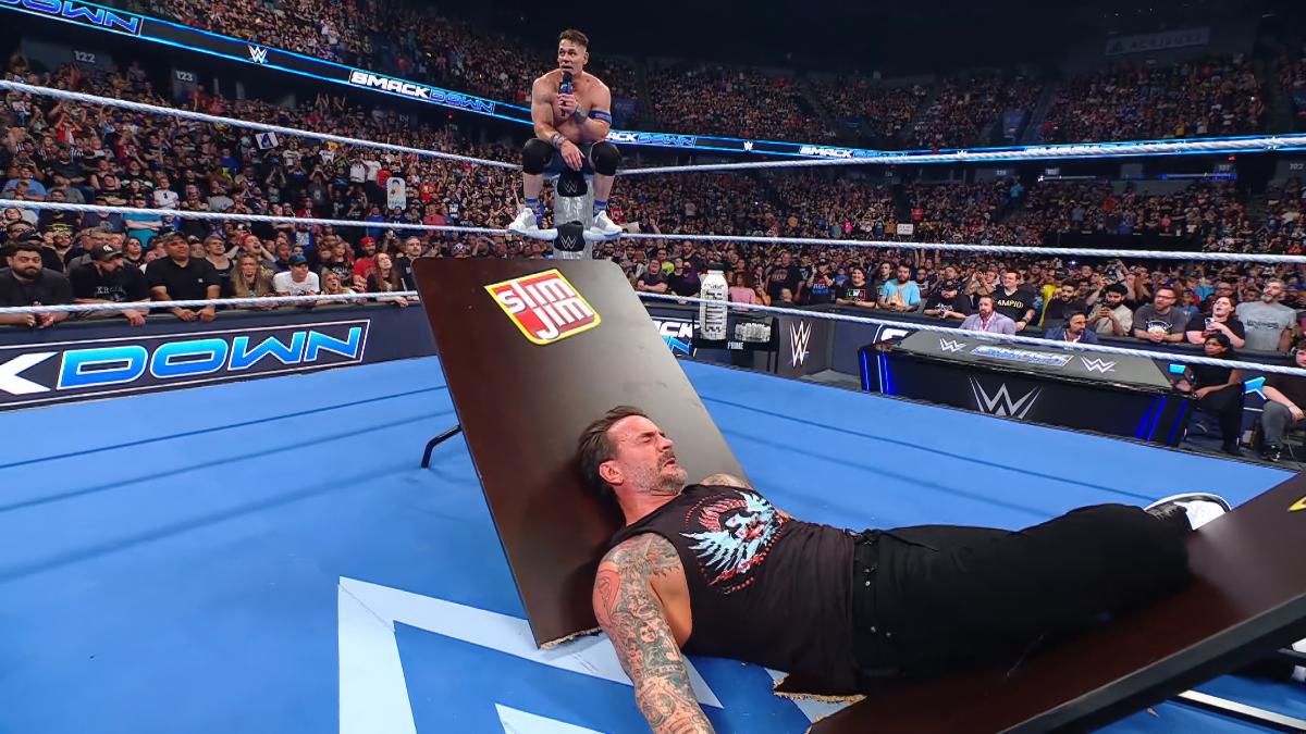 CM Punk lying down in the middle of the ring on a Slim Jim table as John Cena sits on the turnbuckle cutting a promo