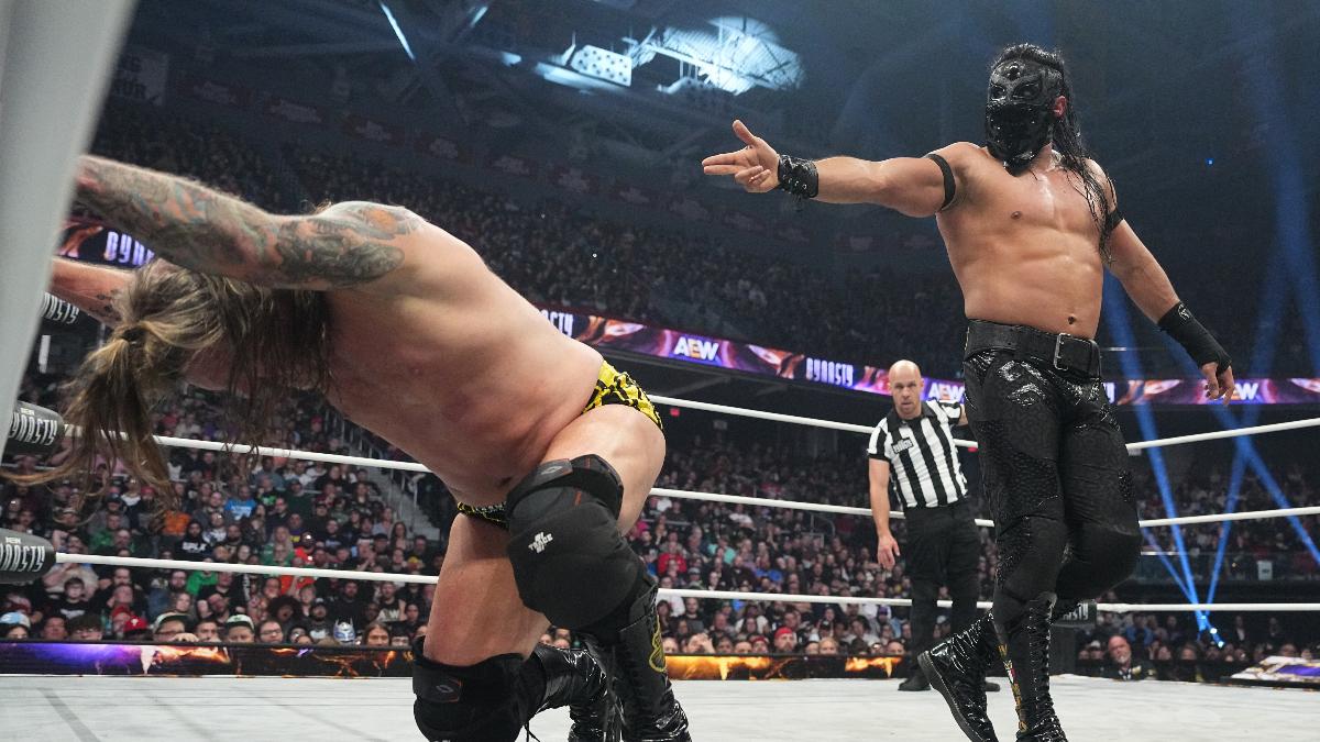 Chris jericho slumped on wrestling ring ropes as Bandido in his lucha mask points a finger gun at his back.