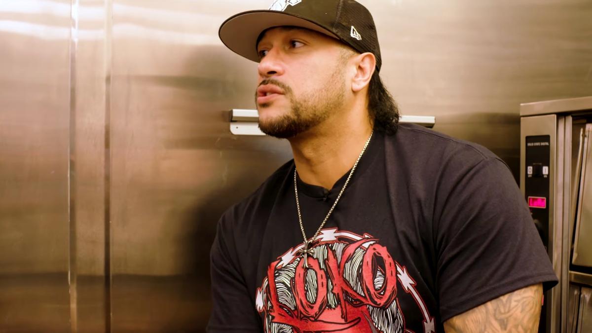 Damian Priest wearing a black t-shirt and cap leaning against a fridge