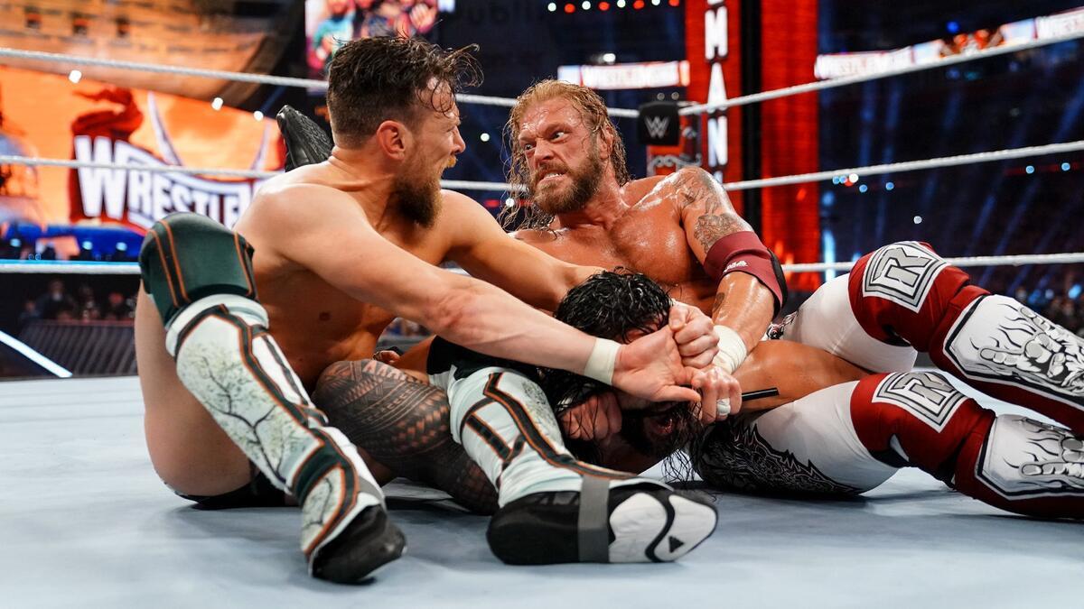 Edge and Daniel Bryan staring angrily at each other as they have Roman Reigns in a submission at WWE WrestleMania 37