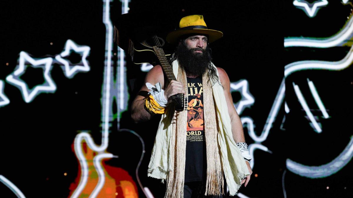 Elijah in a hat,scarf, and cut off tee with a guitar slung over his shoulder, stands on a TNA Wrestling stage