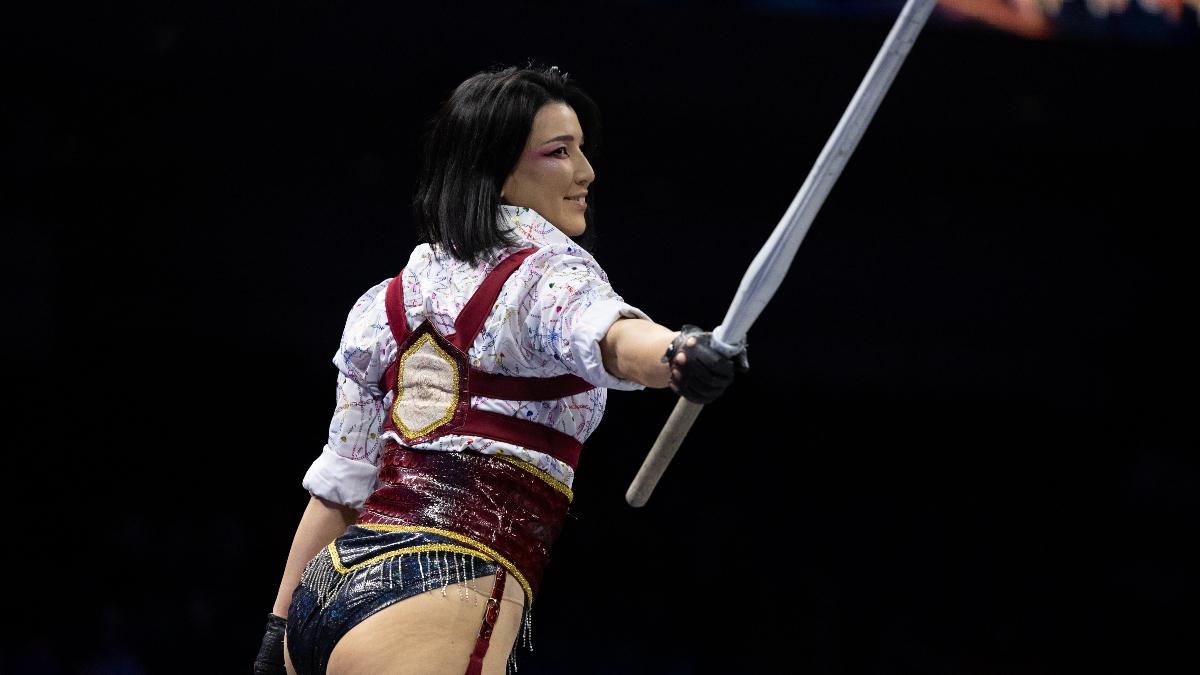 Hikaru Shida smiles with her back half-turned to the camera and holds up a kendo stick