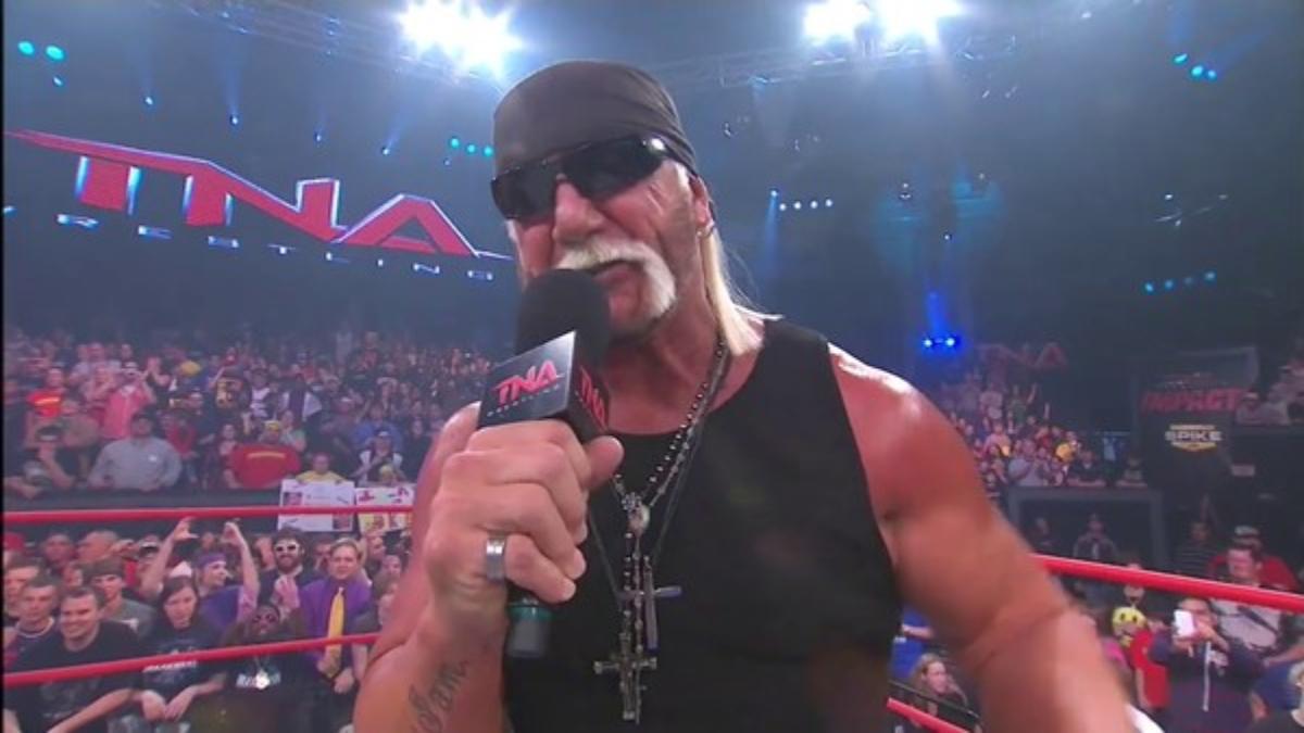 Hulk Hogan in black bandana, sunglasses, and vest speaking on a microphone in a TNA arena