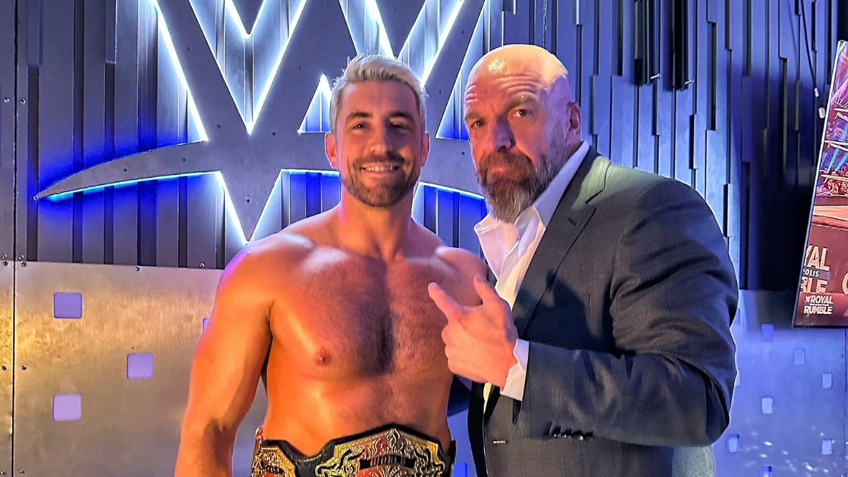 A suited Triple H pointing at Joe Hendry in front of a WWE logo backstage at an event.