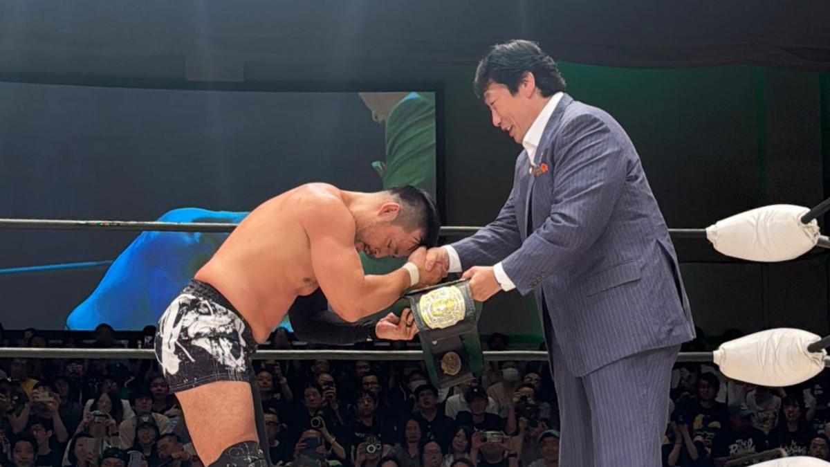 KENTA bows as he receives a championship title belt from a suited Kenta Kobashi in a NOAH ring