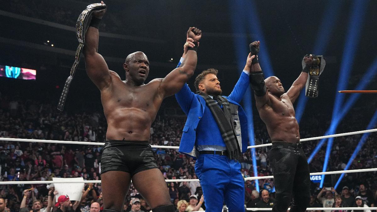 MJF in a blue suit raises the hands of Shelton Benjamin and Bobby Lashley in a wrestling ring, the latter two men holding wrestling championship belts