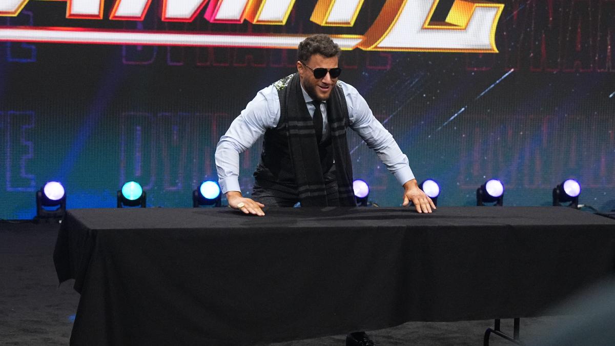 MJF wearing sunglasses while pushing his hands down on a black-blanketed tabled on June 25, 2025 episode of AEW Dynamite