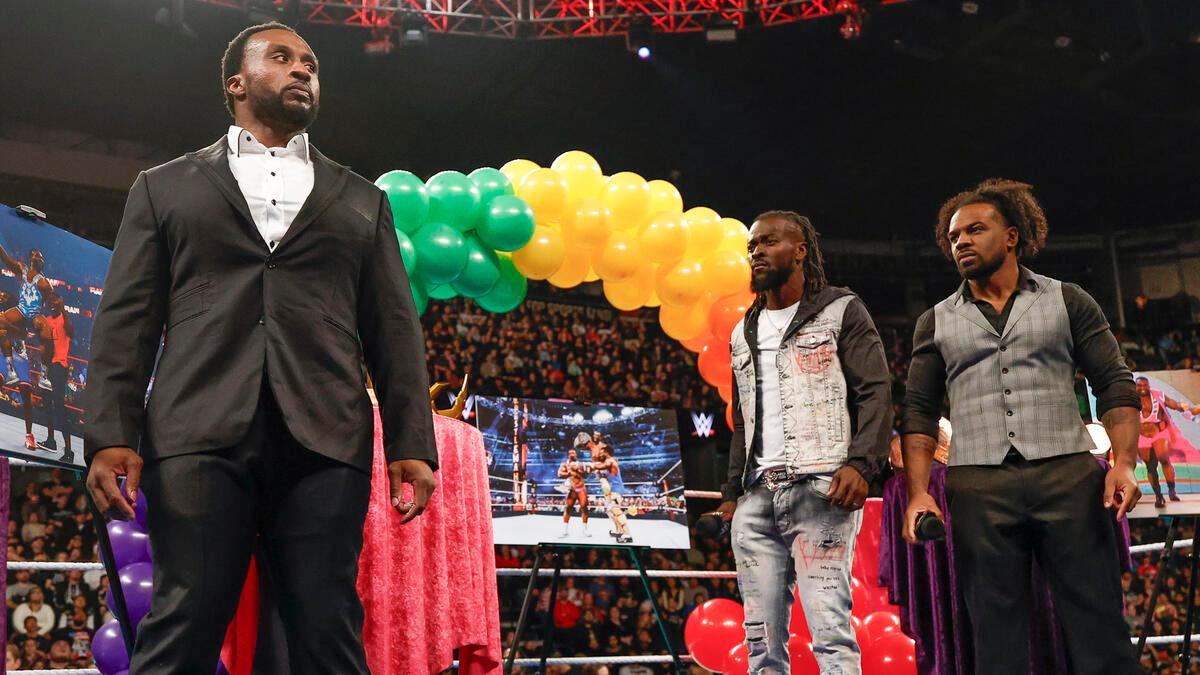 Big E in a suit is stared at by Kofi kingston and Xavier Woods in a wrestling ring with colorful balloons and a framed picture in the background.