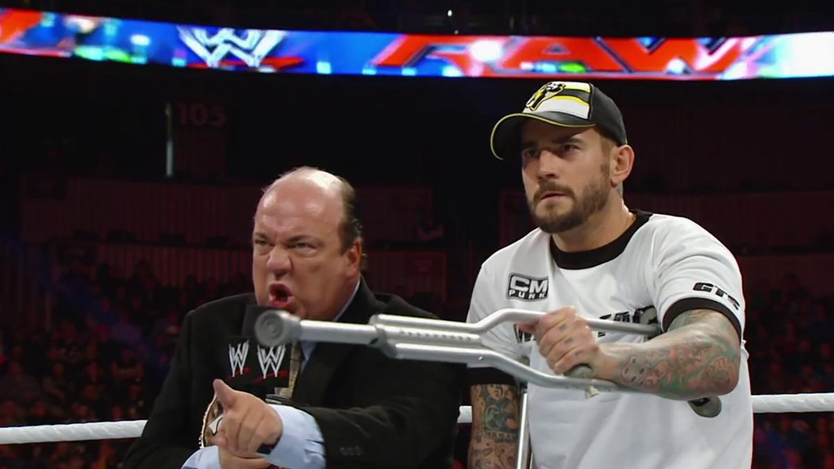 Paul Heyman angrily shouting into a microphone as CM Punk points on crutches 