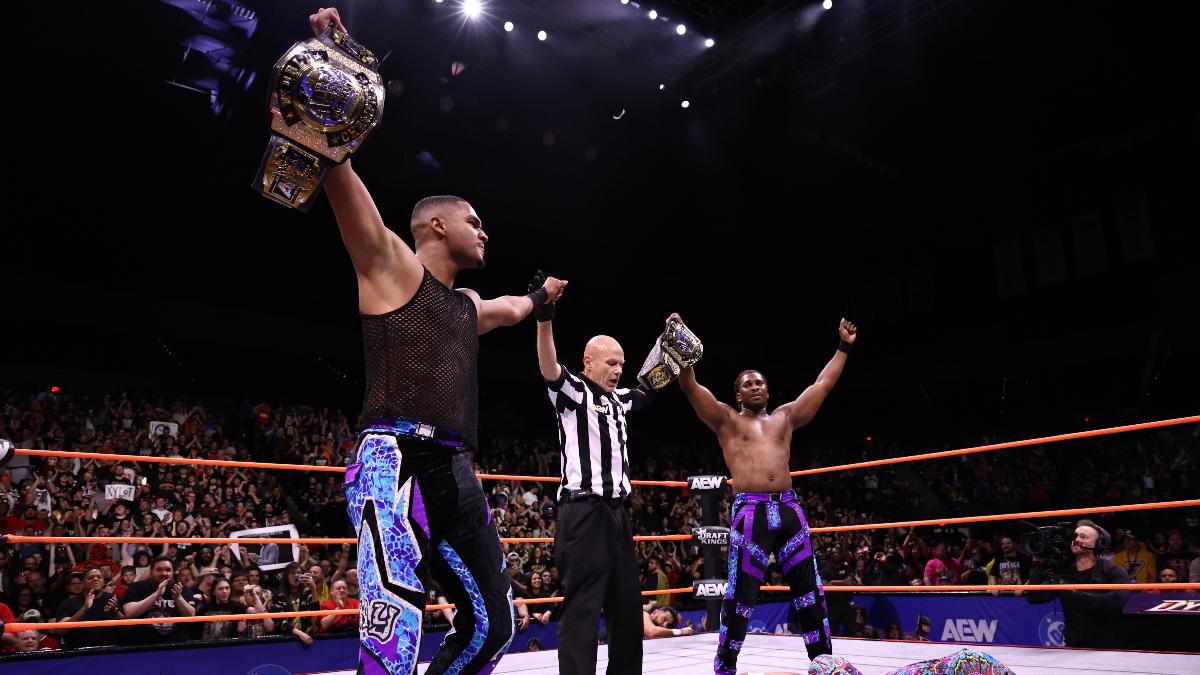 Zay on the left and Quen on the right with referee Rick Knox in the middle hold the AEW Tag Title belts aloft in the ring.