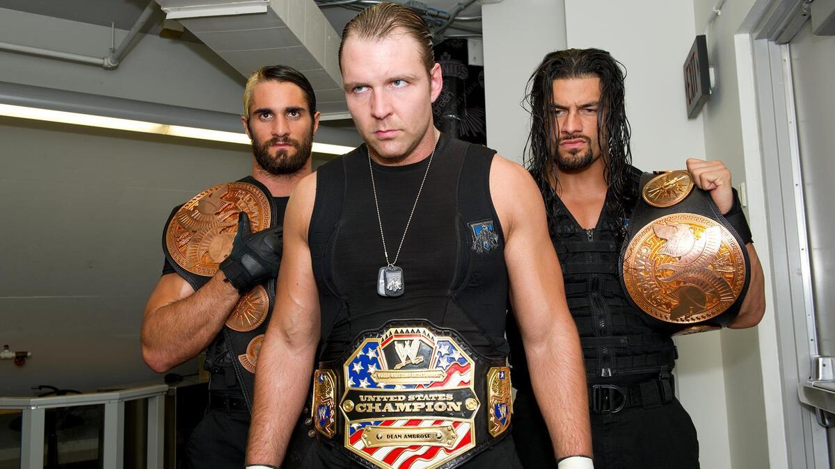 The Shield with Dean Ambrose wearing the WWE United States Title while Roman Reigns and Seth Rollins flank him with the WWE Tag Team Titles