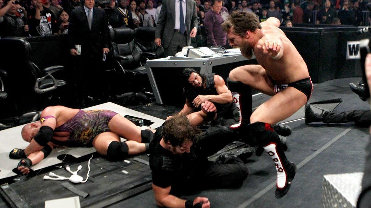 Daniel Bryan kicking Dean Ambrose as Ryback lays down behind them on top of a collapsed announce table