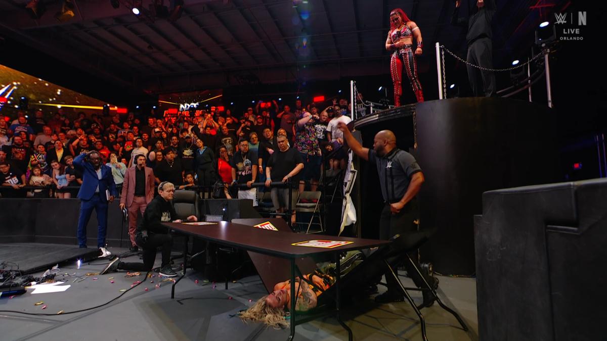 Zaria standing on the platform as Sol Ruca lies on a broken table and commentators Booker T and Vic Joseph look on in shock