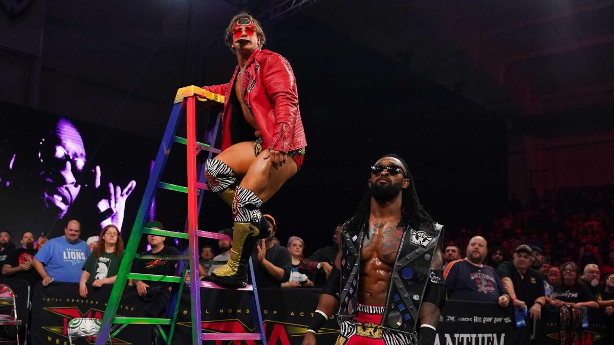 Ace Austin in a red jacket up a colourful ladder with Chris Bey posing on the floor to the right of him