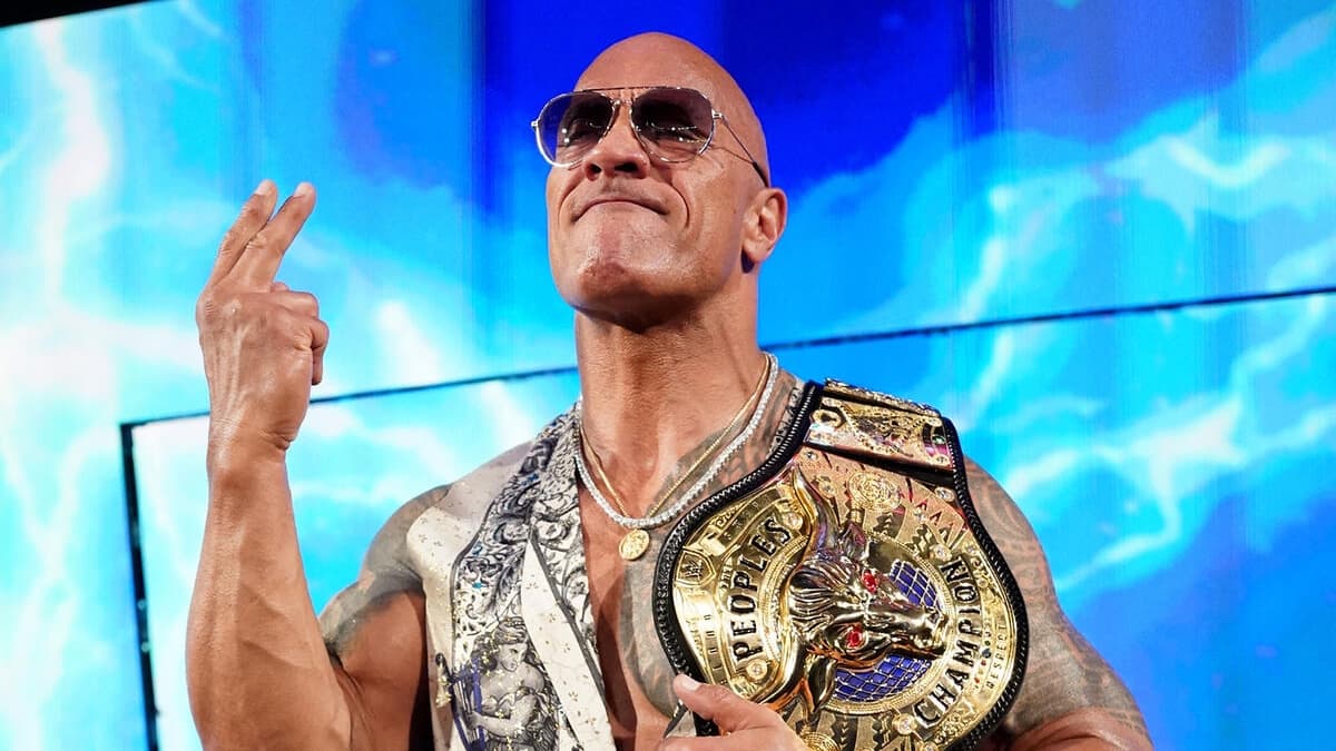 The Rock with his People's Champion belt in a vest holding up two fingers