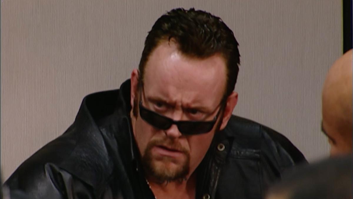 The Undertaker, wearing a leather jacket and with his sunglasses slightly lowered looking disgusted
